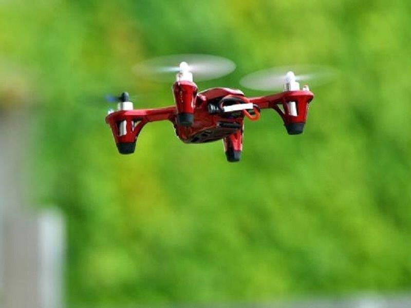 Petit drone rouge en vol stationnaire avec hélices en mouvement sur fond flou de verdure en extérieur.