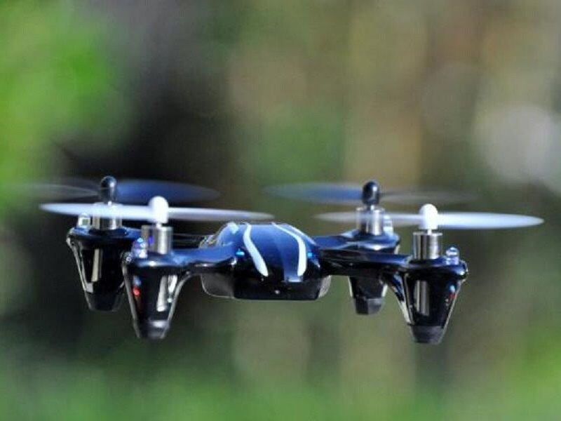 Drone noir et blanc en vol stationnaire avec ses hélices en mouvement, fond flou de verdure en arrière-plan.