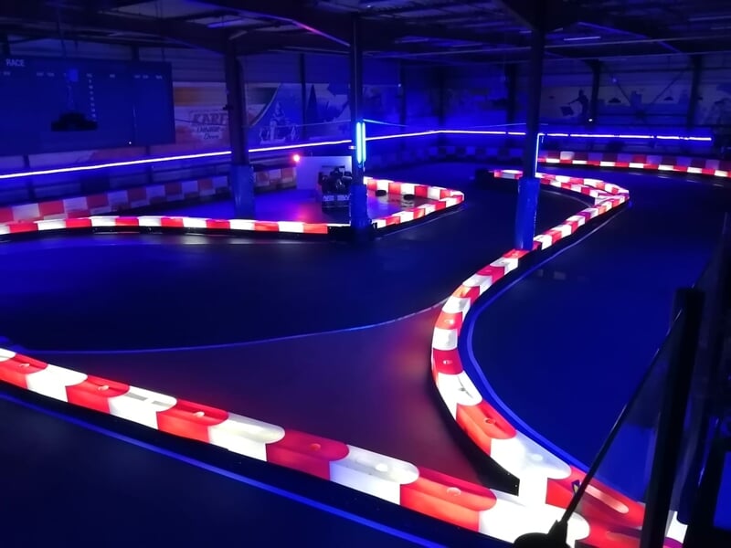 Piste de karting intérieure avec barrières rouges et blanches sous éclairage bleu, virages serrés visibles.