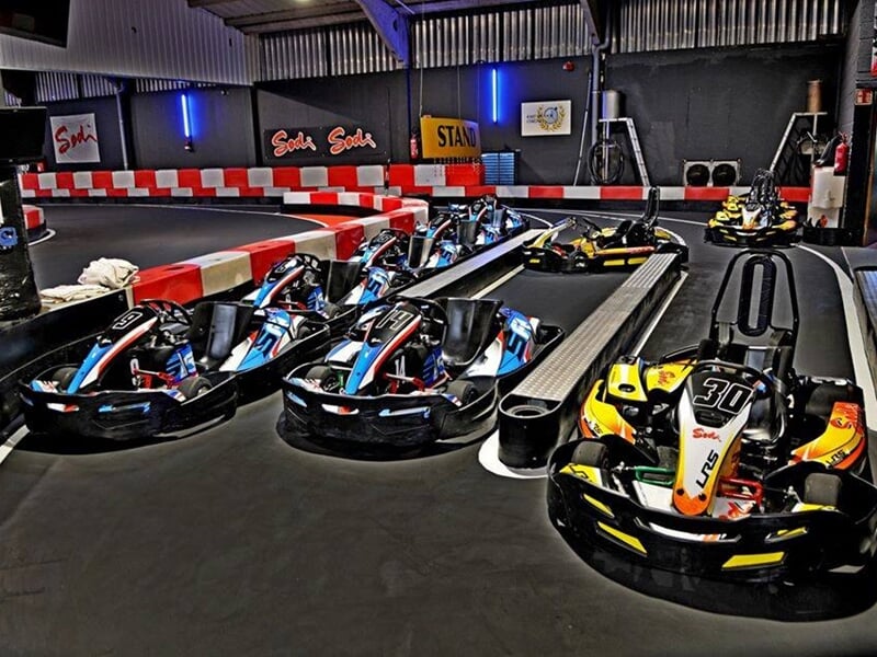 Cinq karts alignés dans un garage ou zone de départ, avec barrières rouges et blanches en arrière-plan.