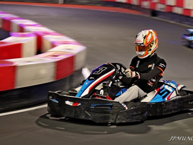 Pilote en karting en pleine action sur une piste intérieure, portant un casque orange et une combinaison noire.