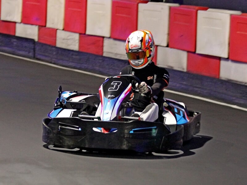 Pilote seul en karting sur piste intérieure, en virage, avec casque blanc et combinaison noire.
