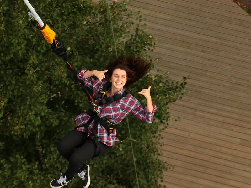 Jeune femme souriante en train de faire un saut à l'élastique, suspendue au-dessus d'un sol en bois et de la végétation.
