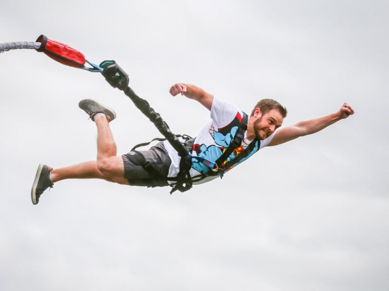 Homme en tenue décontractée effectuant un saut à l'élastique, bras tendus, avec un ciel nuageux en arrière-plan.
