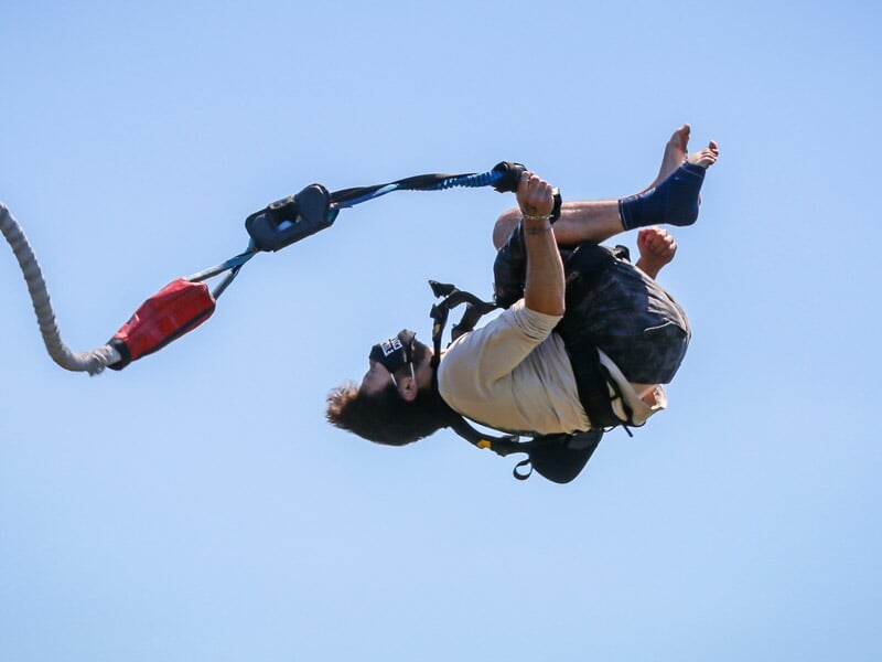 Personne en combinaison et harnais effectuant un saut à l'élastique, suspendue dans les airs avec un ciel bleu clair.