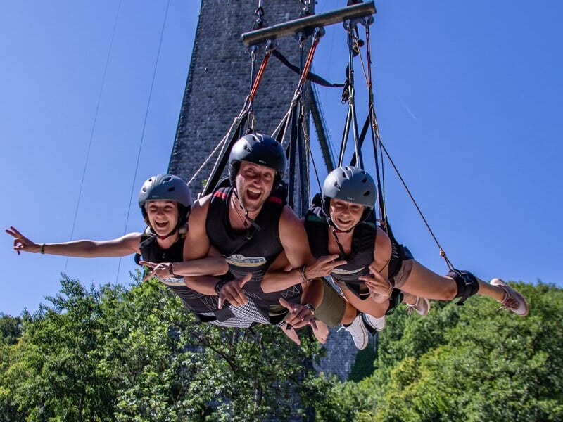 Trois personnes souriantes en train de faire un saut en balançoire géante côte à côte, avec un grand viaduc en arrière-plan