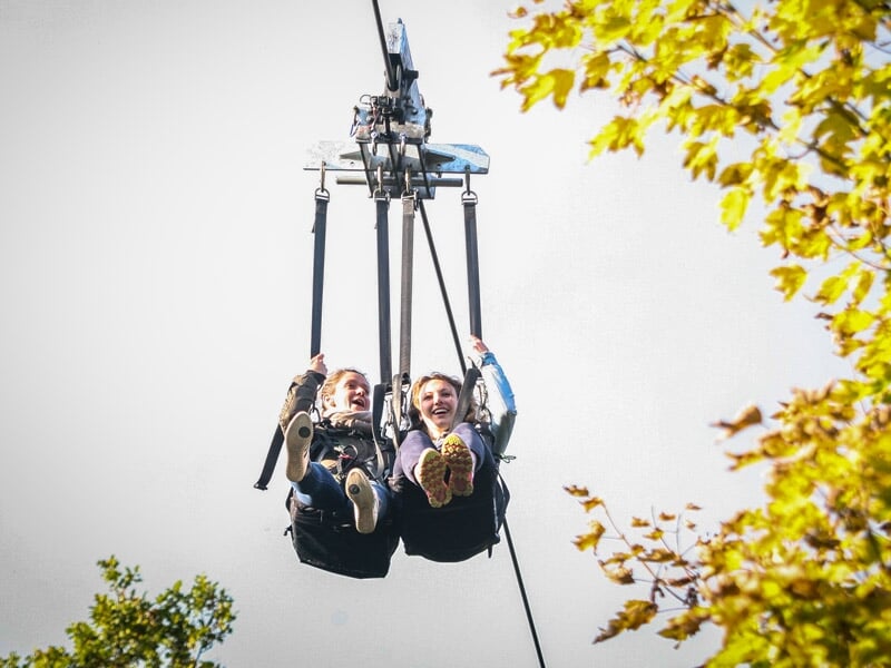 Deux femmes souriantes en train de faire un saut à l'élastique ensemble, suspendues à un harnais au-dessus des arbres en automne