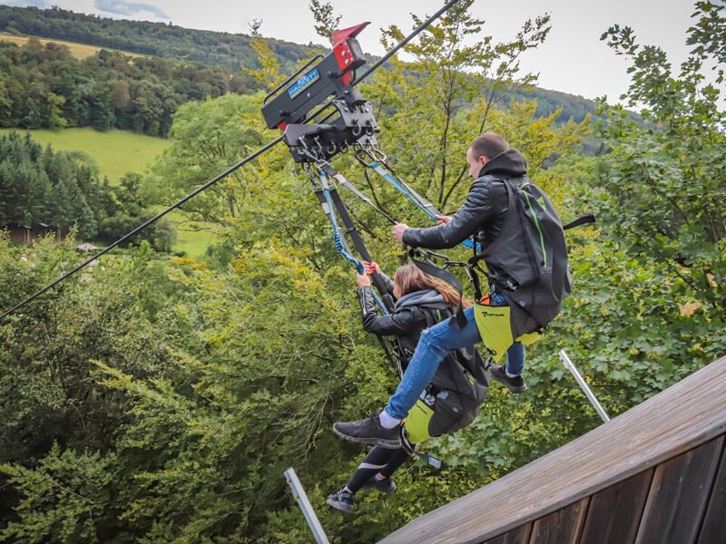 Un homme et une femme attachés à un système de câbles pour un saut à l'élastique, se préparant à sauter au-dessus d'une forêt luxuriante