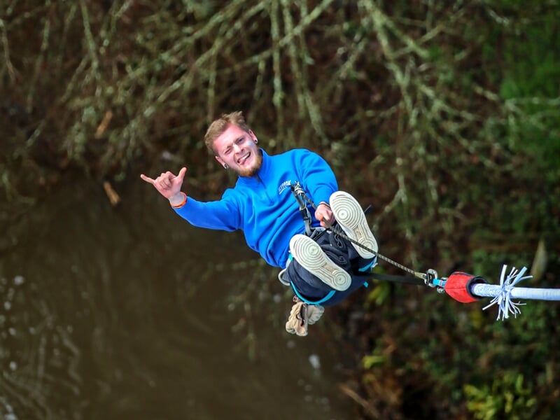 Un homme avec un haut bleu qui fait un saut à l'élastique au-dessus d'un plan d'eau, en faisant un signe de la main