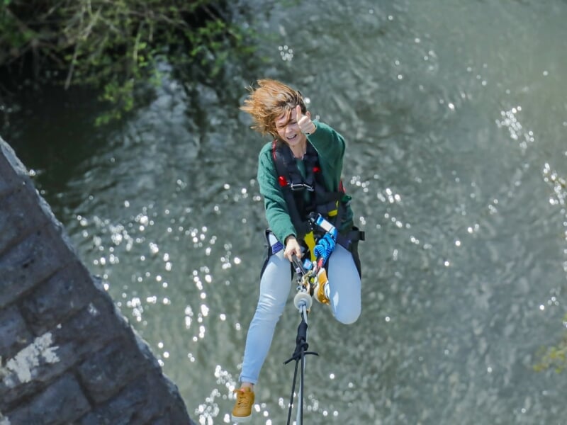 Femme avec un pull vert et des chaussures jaunes, accrochée à une tyrolienne au-dessus d'une rivière, exprimant la joie.