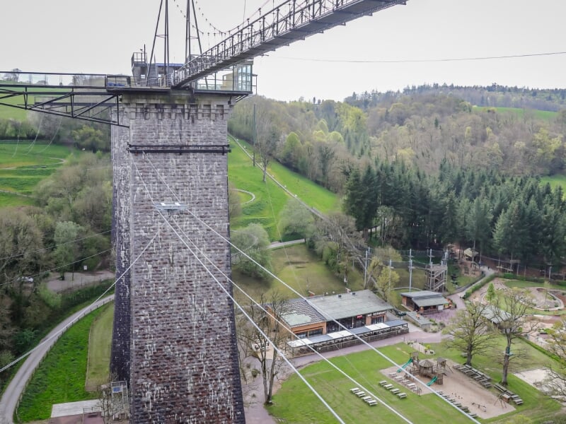 Vue panoramique d'un pont en pierre avec une structure métallique, traversant une vallée verdoyante, avec des bâtiments et des aires de jeux en dessous.