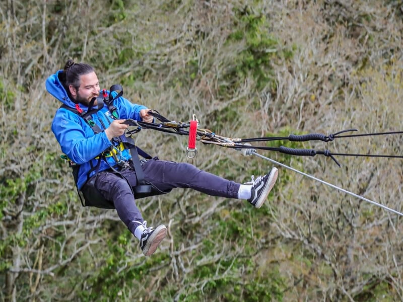 Homme avec une barbe et un sweat-shirt bleu, attaché à un harnais de tyrolienne, glissant au-dessus d'une forêt.