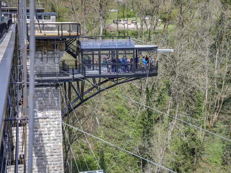 Groupe de personnes sur une plateforme d'observation métallique avec un toit en verre, surplombant une vallée boisée.
