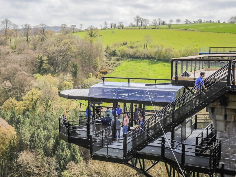 Plateforme d'observation avec des visiteurs, entourée de collines verdoyantes et de forêts, accessible par un escalier métallique.