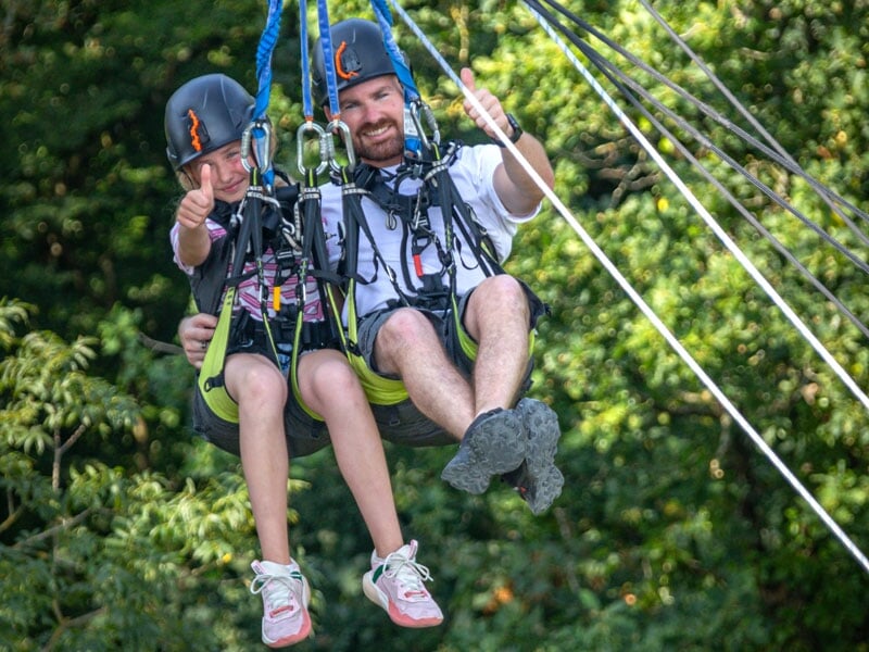 Un père et sa fille, casqués, exécutent un saut pendulaire ensemble, tous deux affichant un pouce levé en signe d'excitation