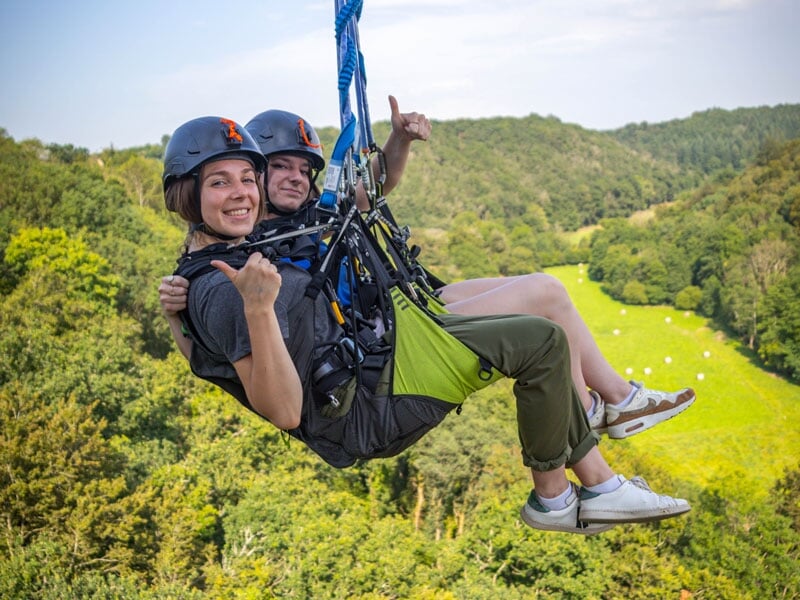 Gros plan sur deux personnes souriantes lors de leur saut pendulaire, avec en arrière-plan un paysage vallonné et verdoyant