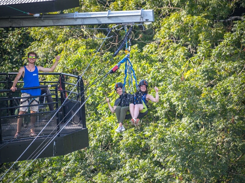 Un duo de participants est suspendu au-dessus de la forêt, tandis qu'un instructeur les salue depuis une plateforme métallique