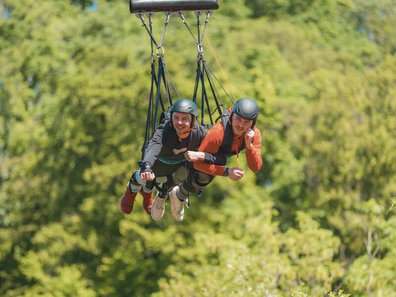 Deux personnes en combinaison et casque effectuent un saut élastique en tandem au-dessus d'une forêt verdoyante sous un ciel clair.