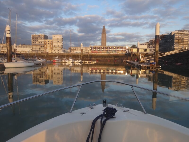 Vue depuis un bateau dans un port calme avec des voiliers et des bâtiments sous un ciel nuageux.