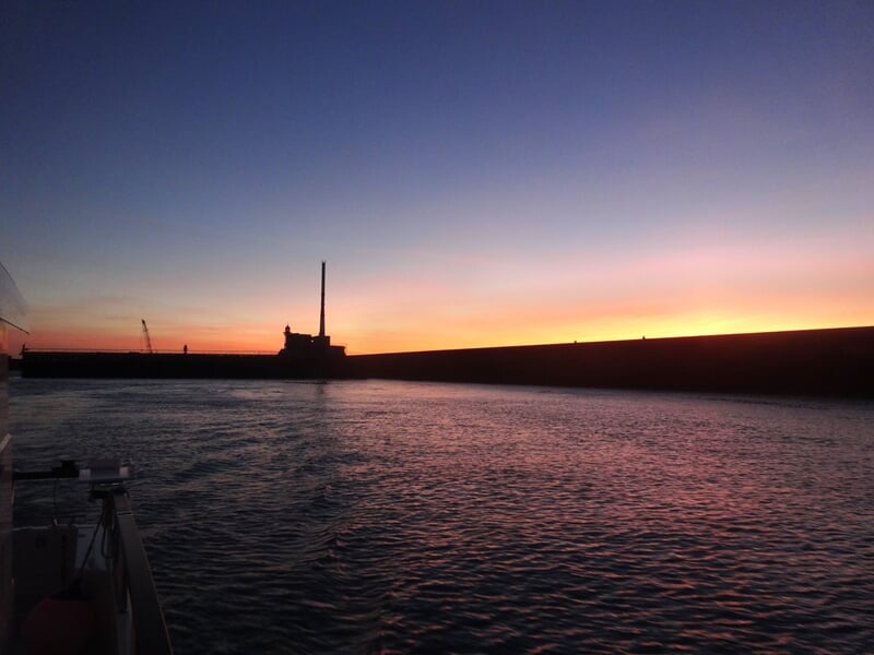 Coucher de soleil sur un port avec un grand mur de quai et un ciel aux teintes roses et violettes.