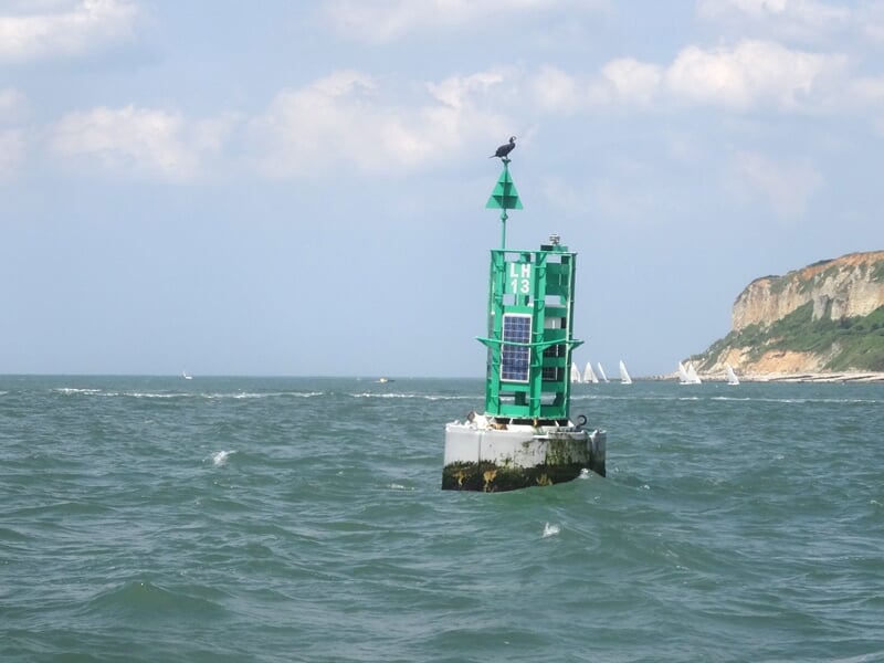 Bouée verte sur une mer agitée avec des falaises blanches et plusieurs voiliers au loin.