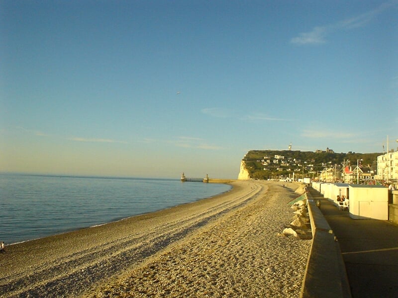 Plage de galets avec une promenade et des falaises blanches sous un ciel clair en fin de journée.