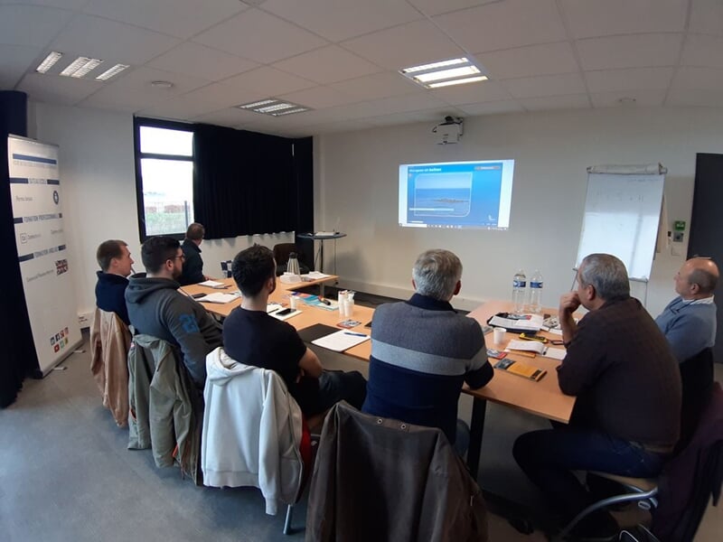Salle de formation avec plusieurs personnes assises, regardant un écran dans une pièce lumineuse.