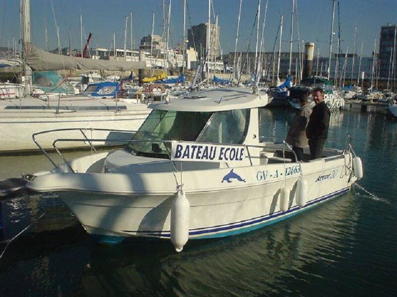 Bateau école blanc amarré dans un port avec deux personnes debout à bord, voiliers en arrière-plan sous un ciel clair.