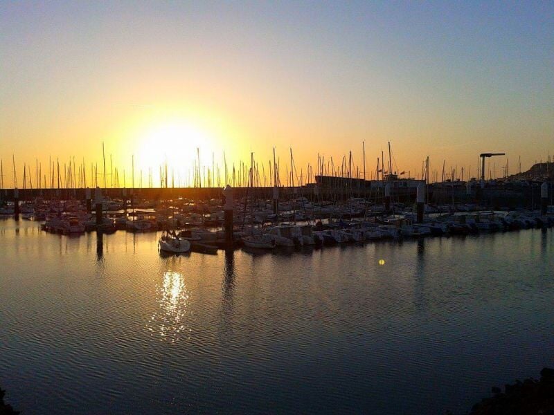 Coucher de soleil sur un port avec de nombreux bateaux à voile amarrés et reflet doré sur l'eau.