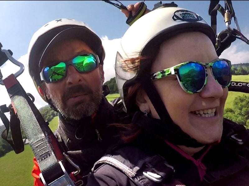 Selfie de deux personnes en parapente, l'homme avec une barbe portant un casque et des lunettes de soleil vertes, la femme souriant avec des lunettes de soleil colorées