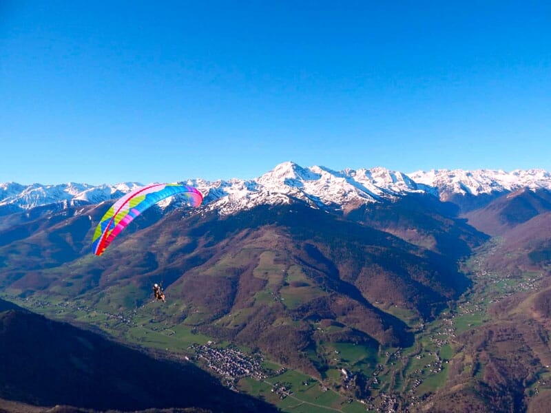 Parapente coloré volant au-dessus d'un paysage montagneux verdoyant avec des sommets enneigés en arrière-plan sous un ciel bleu clair
