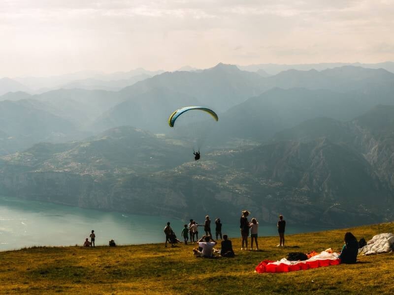 Un parapentiste survole un paysage montagneux avec un lac, observé par un groupe de spectateurs sur une colline.