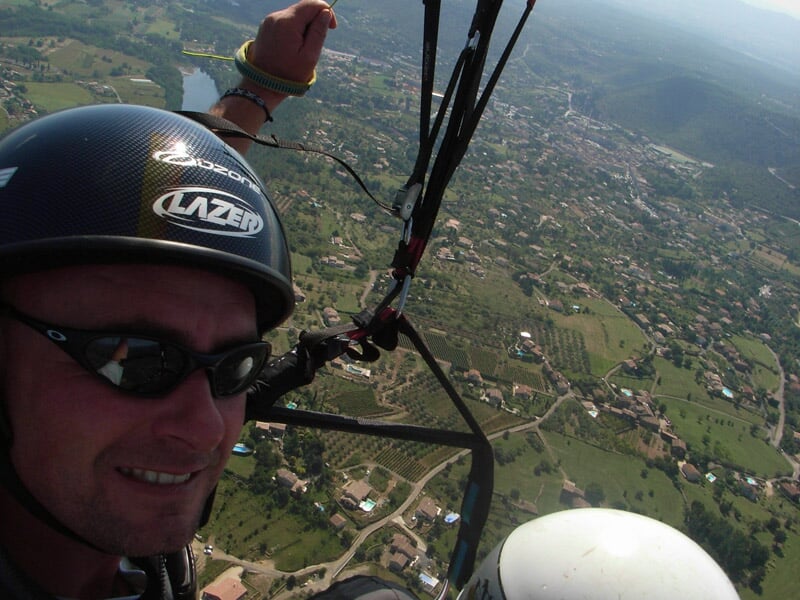 Selfie d’un parapentiste en plein vol au-dessus d’une campagne parsemée de maisons et d’arbres, portant un casque noir et des lunettes de soleil.