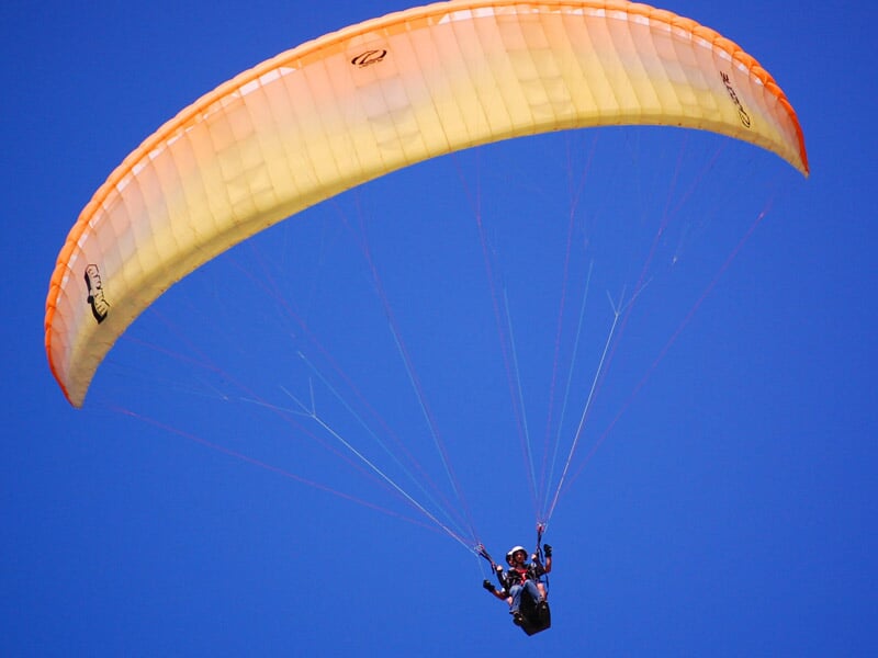 Parapente jaune en plein ciel bleu, avec un pilote visible sous la voile, suspendu par des suspentes.
