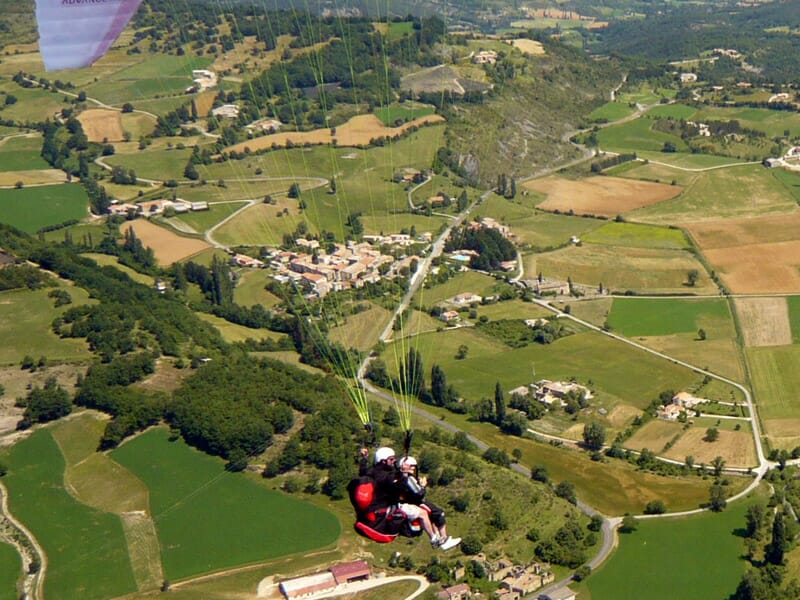 Deux parapentistes en vol au-dessus d'une campagne composée de champs, routes sinueuses et petits villages.