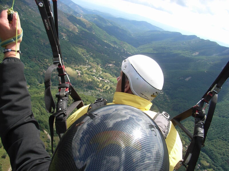 Vue aérienne d'un parapentiste survolant une vallée verdoyante, avec des montagnes en arrière-plan et un casque blanc visible au premier plan.