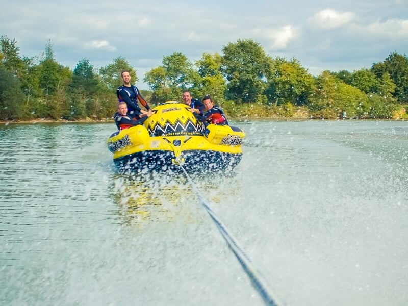 Quatre personnes sur une bouée tractée jaune sur un lac, tirée par un bateau en mouvement.