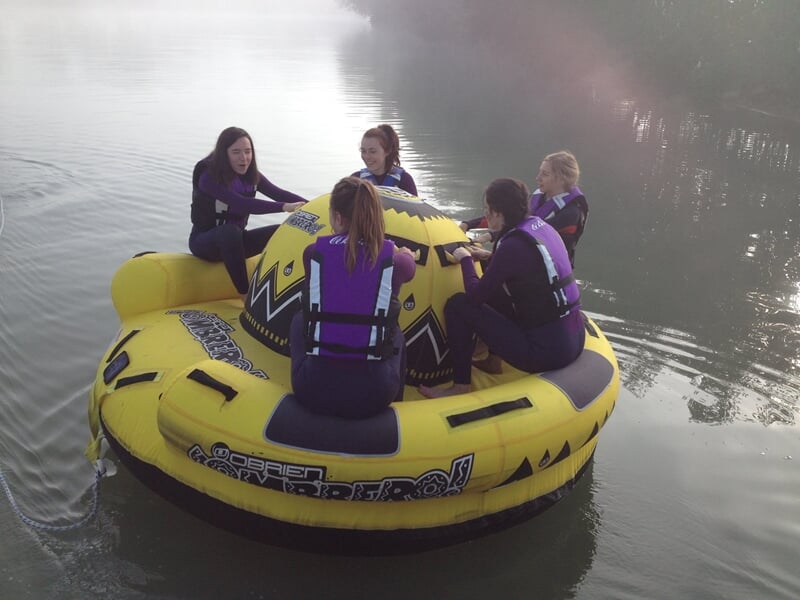 Cinq personnes en gilets de sauvetage sont assises sur une bouée tractée jaune sur un lac calme.