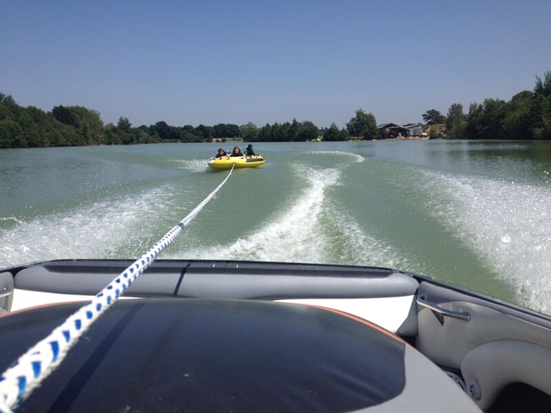 Vue depuis un bateau sur un lac, une bouée tractée jaune avec plusieurs personnes est tirée à grande vitesse.