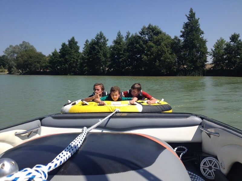 Trois enfants en gilets de sauvetage sur une bouée tractée jaune, vus depuis un bateau sur un lac.