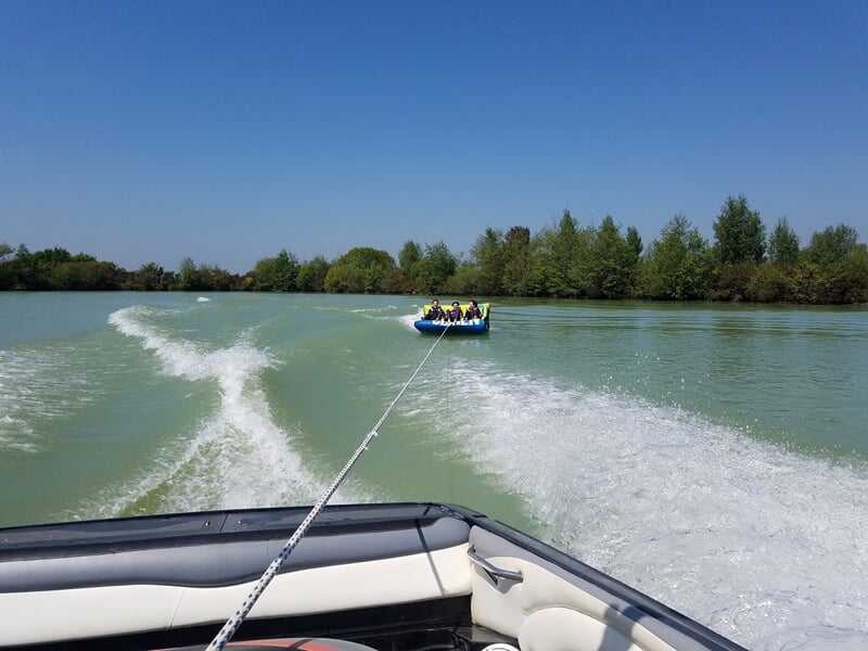 Une bouée tractée bleue avec plusieurs personnes est tirée sur un lac, vue depuis un bateau en mouvement.