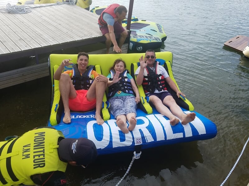 Trois jeunes assis sur une bouée tractée bleue sourient, attachés à un bateau sur un lac calme.