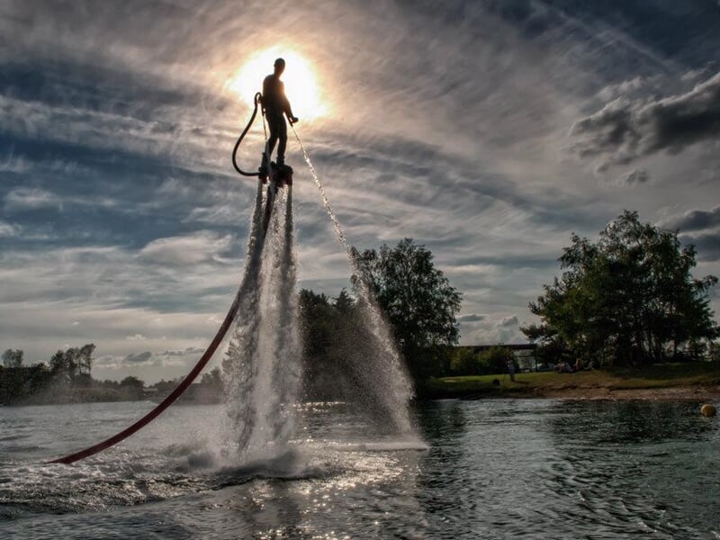 Un homme en combinaison noire utilise un flyboard, propulsé par de l'eau, sur un lac sous un ciel nuageux.