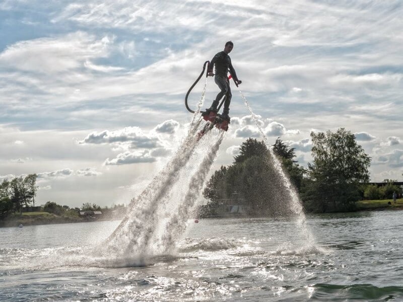 Un homme en flyboard s'élève au-dessus de l'eau, entouré de jets d'eau puissants sous un ciel nuageux.