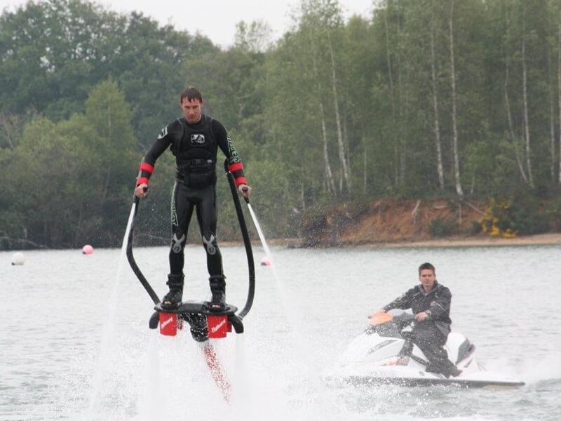 Un homme en combinaison noire fait du flyboard sur un lac, avec un jetski à proximité.
