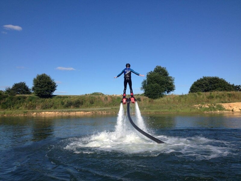 Un homme en flyboard s'élève au-dessus d'un lac, entouré d'arbres verts sous un ciel bleu.