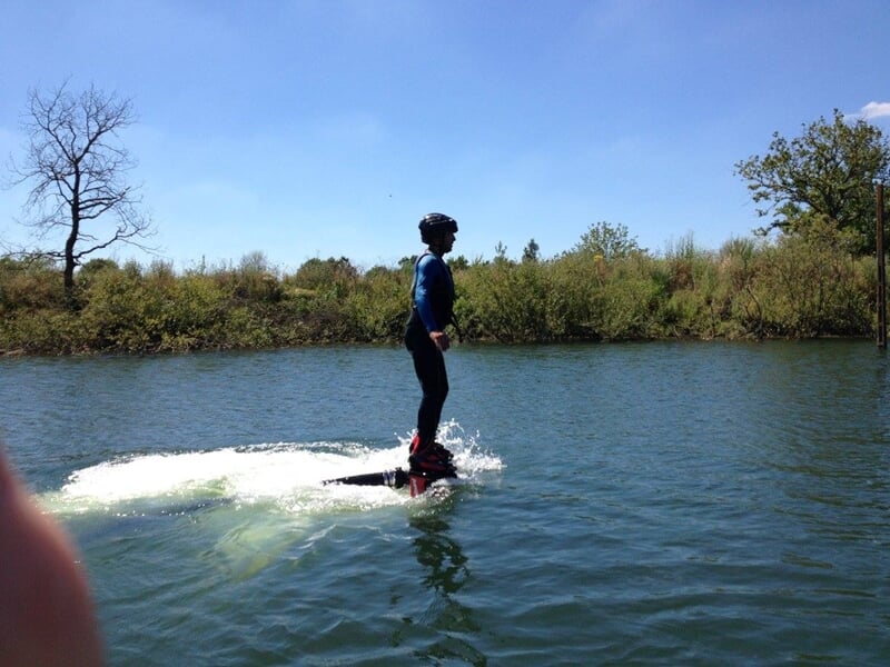 Un homme en flyboard sur un lac calme, les bras écartés, sous un ciel bleu avec quelques nuages.