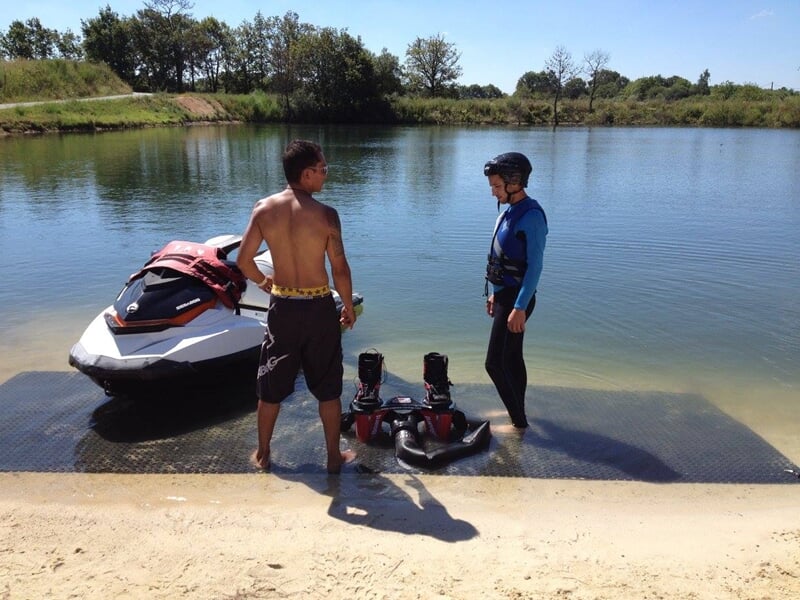 Deux hommes discutent au bord d'un lac, avec un jetski et un équipement de flyboard posés sur le sol.