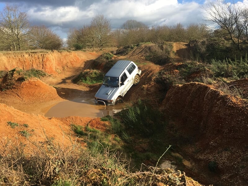 Un 4x4 blanc franchit un obstacle boueux dans un terrain accidenté en pleine nature.