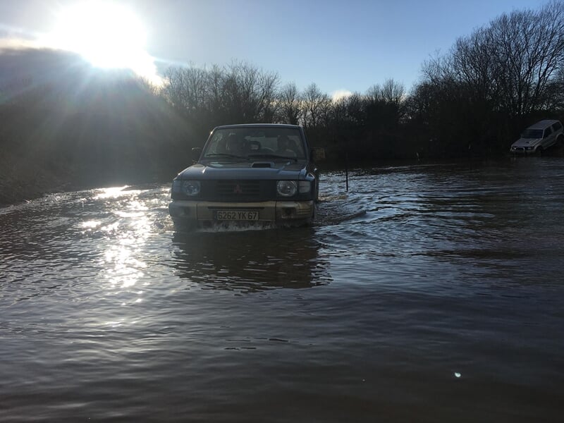 Un 4x4 noir traverse une rivière peu profonde dans un paysage boisé au coucher du soleil.